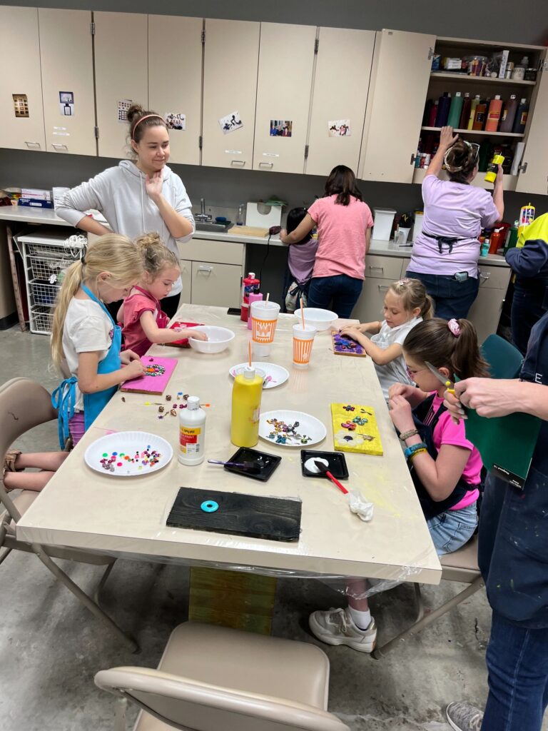 A group of artists at a table decorating boards with gems and glitter, with paint bottles, cups and a finished dark board decorated with a blue gem flower.