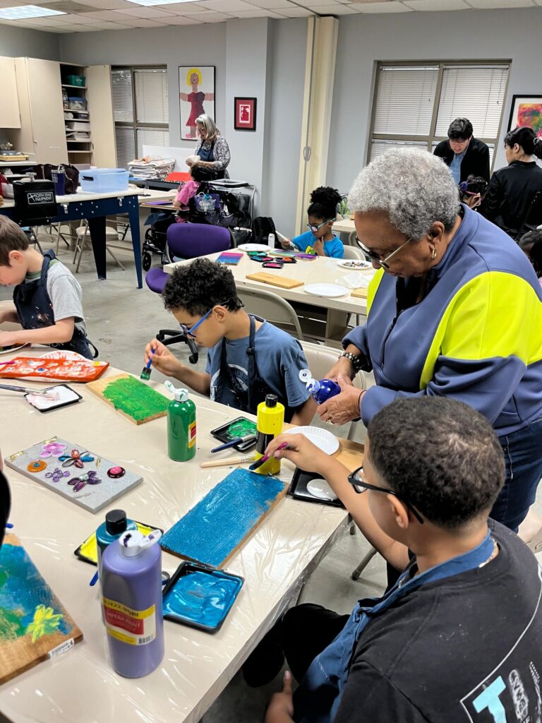 A volunteer helps an artist paint a wooden board blue, with a finished piece decorated with pressed flowers nearby and paint trays set out on the table.