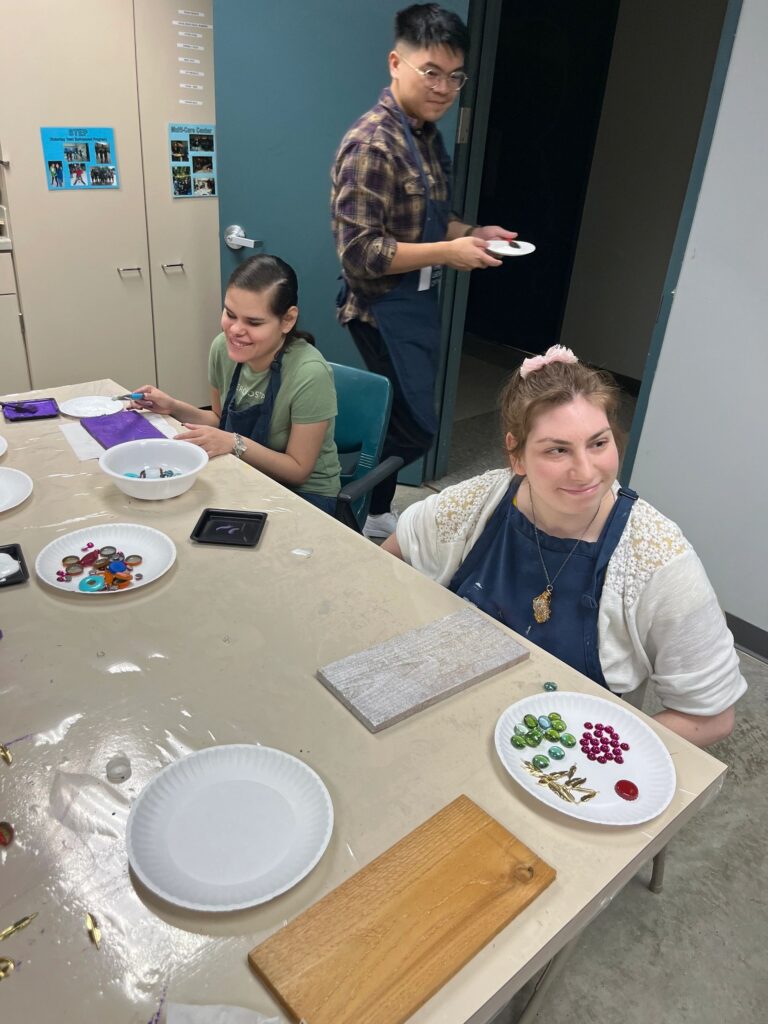Two artists at a table, one arranging glass gems on a plate, a volunteer nearby holding a small plate of decorations.