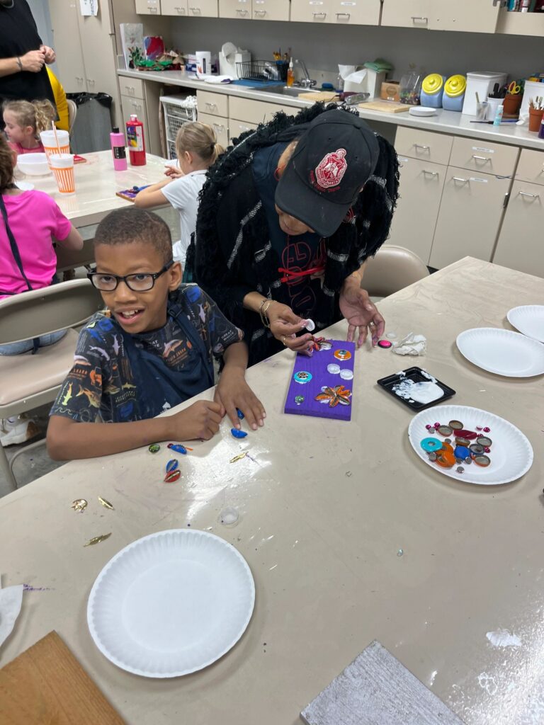 A volunteer helps an artist press bottle caps and gems onto a small purple board, with a plate of extra decorations on the table.