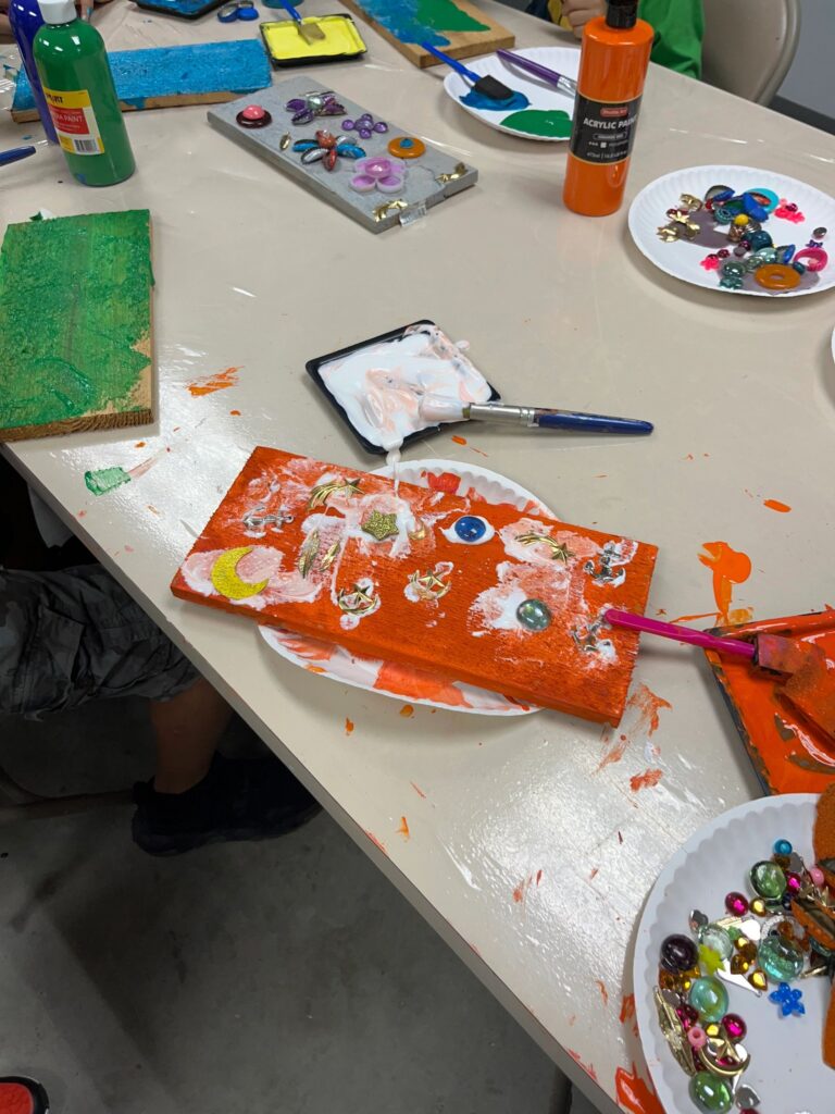 An orange-painted board decorated with glass gems, a crescent moon charm and small gold star charms, with a paintbrush and glue tray resting beside it.
