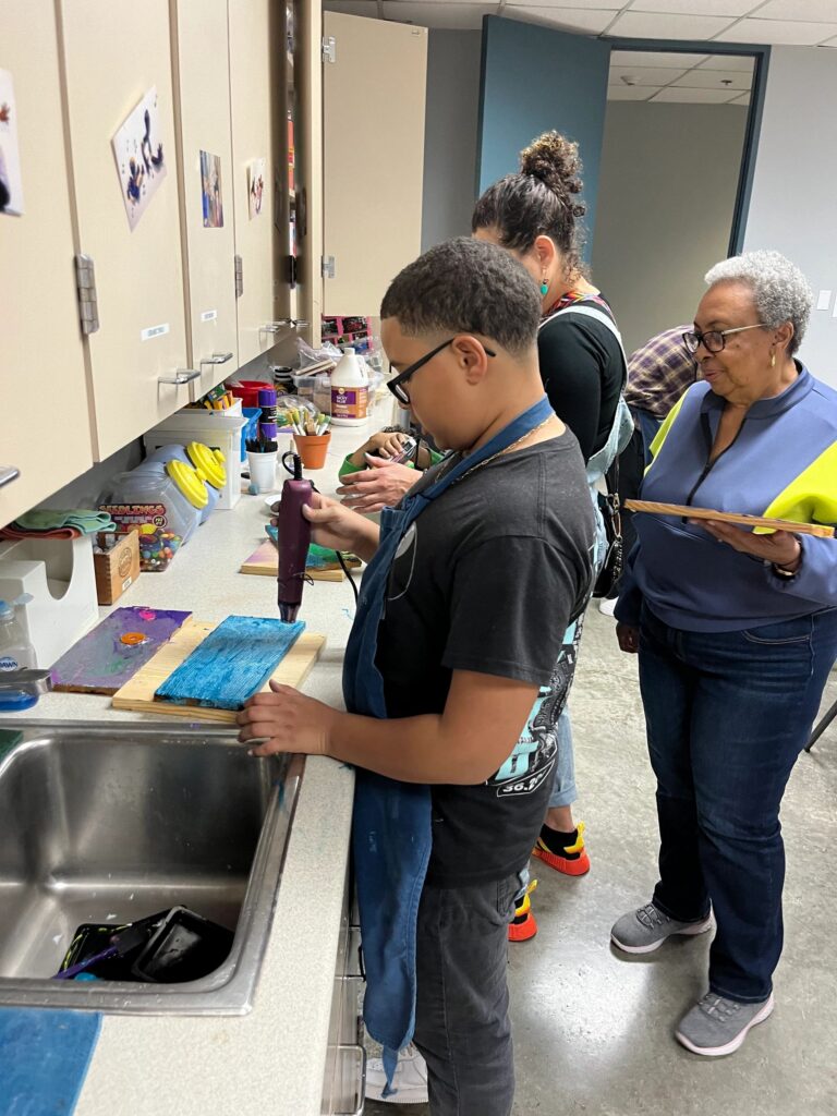 A wide view of the studio, with artists and volunteers gathered around several tables decorating boards with paint and small charms.