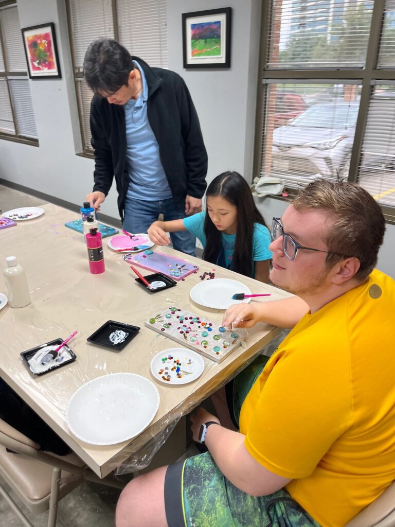A volunteer helps an artist dry a freshly painted blue board with a heat tool at the sink counter, while another volunteer looks on holding a wooden board.