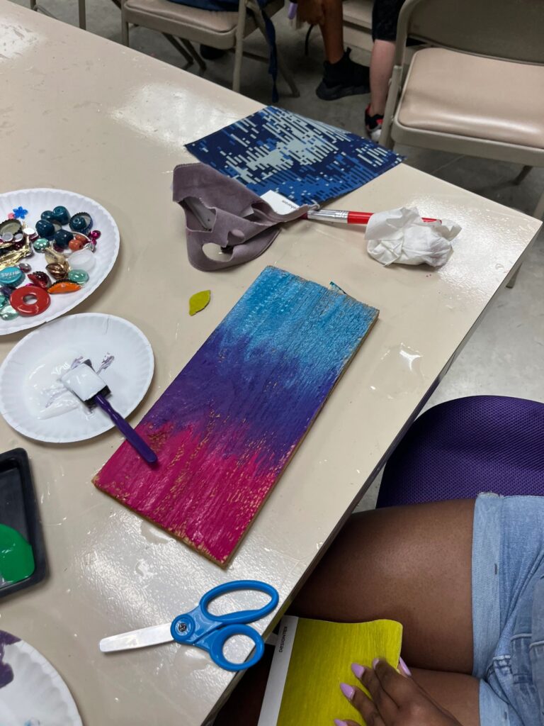 A volunteer helps an artist decorate a purple board with glass gems, with paint bottles, bowls of beads and a finished piece nearby on the table.