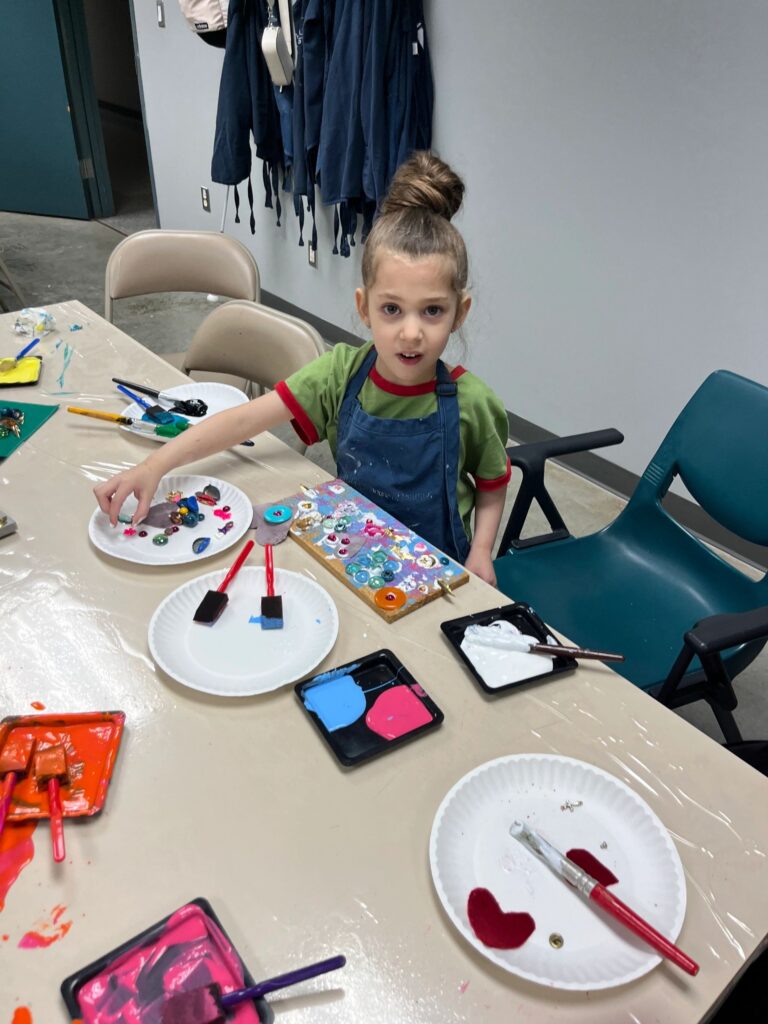 An artist presses paint and small charms onto a board decorated with buttons and gems, with paper plates of paint in red and pink set out beside them.