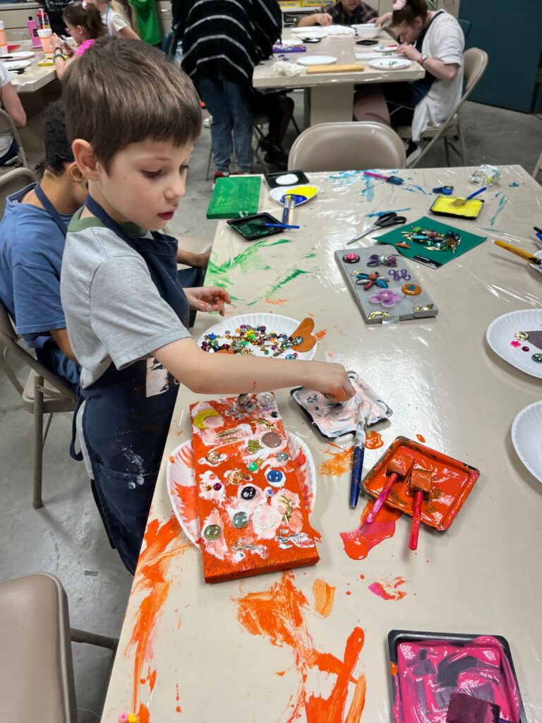 An artist decorates an orange-painted board with buttons and gems, pressing them into wet paint with a foam brush and glue tray on the table.