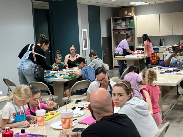 A wide view of the studio, with artists at several tables decorating boards while volunteers set up more materials at a table in the background.