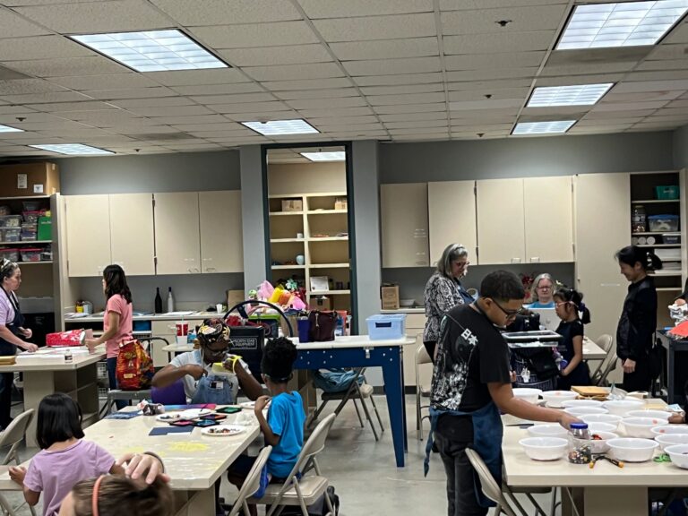 A wide view of the studio from across the room, with artists and volunteers at tables and shelves of supplies along the back wall.