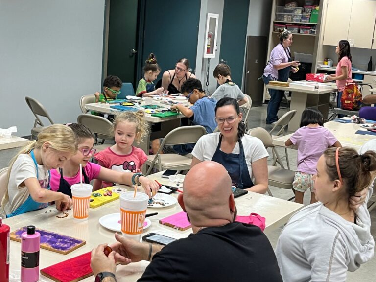 A group of artists and volunteers at a table, several arranging gems and glitter on painted boards, with cups and paint bottles spread across the tabletop.