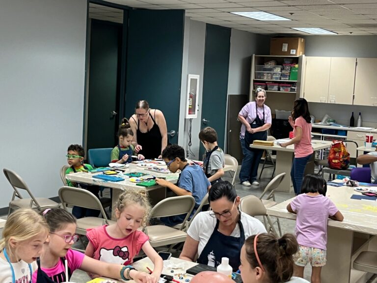 A wide view of the studio, with several groups of artists and volunteers working at tables across the room, one board visible with blue and green paint.