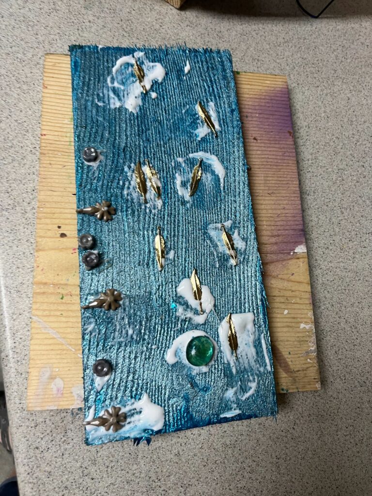 A textured turquoise panel resting on a workbench, its surface combed into fine ridges and decorated with brass feather charms, small metal flowers and a single green glass gem set in white modelling paste.