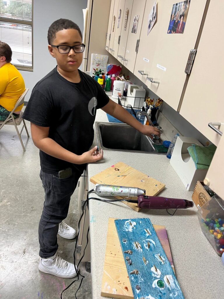 An artist dries a finished turquoise board with feather charms and a green glass gem using a heat tool at the sink counter, with a bin of glass marbles nearby.