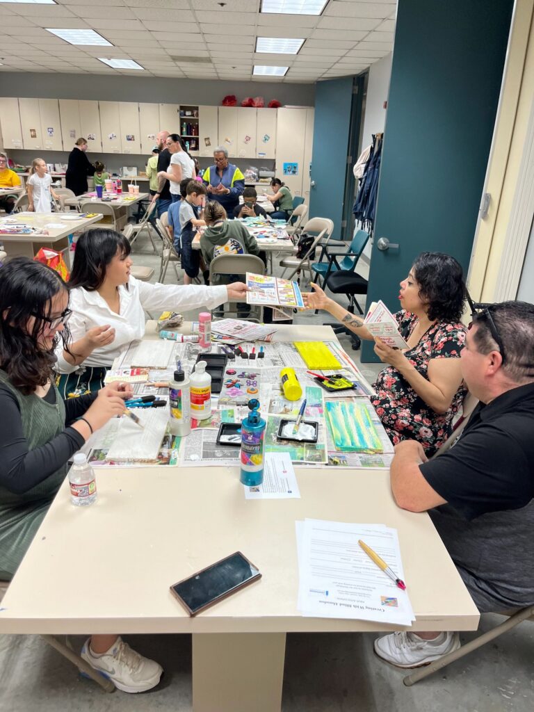 A group of artists and volunteers seated around a table covered in newspaper, sharing a printed flyer while a painted board and craft supplies sit among glue bottles and paint trays.