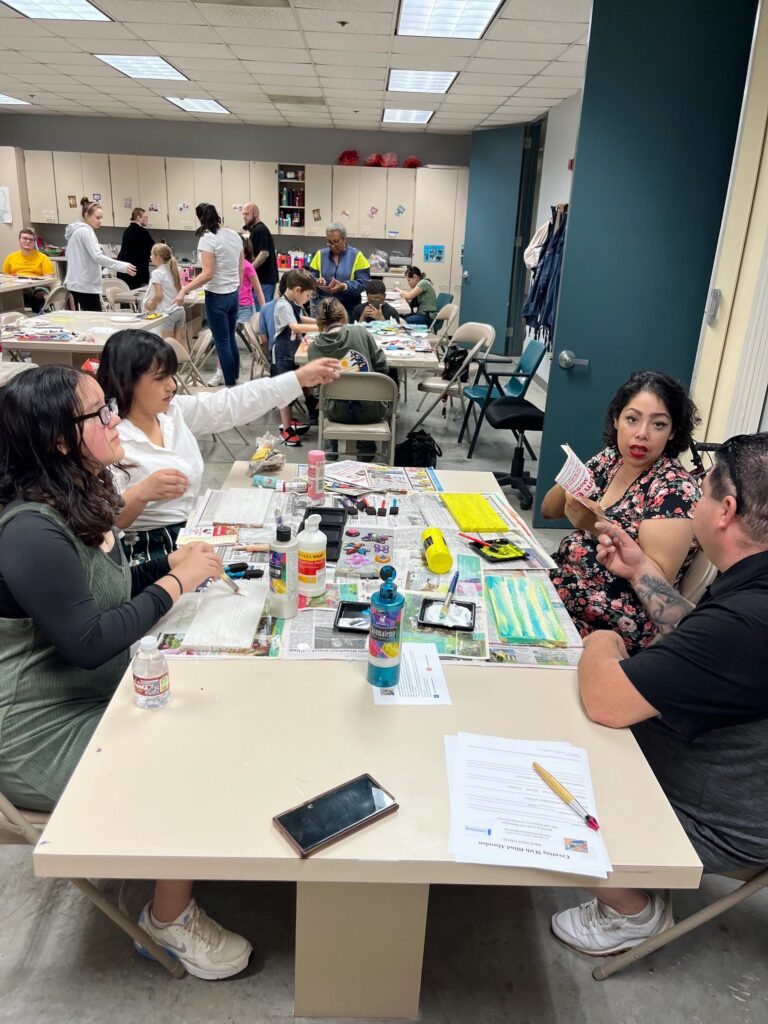 The same group at the table from a slightly different angle, with a volunteer passing a flyer across the table and craft supplies spread over the newspaper covering.
