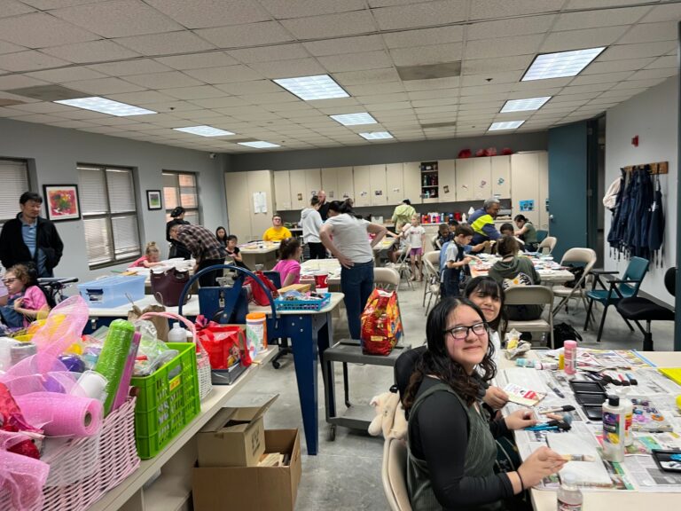 A wide view of the studio, with artists at several tables and bins of ribbon and craft supplies lined up along a side table in the foreground.