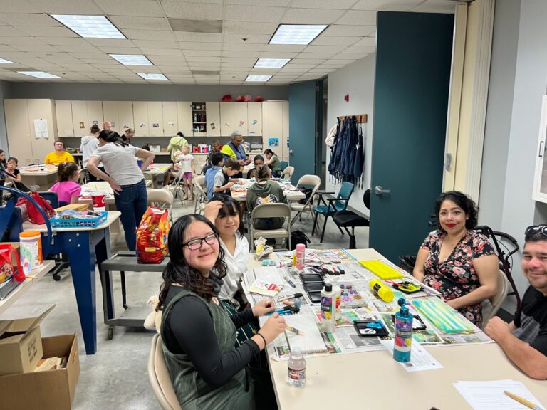 An artist smiles at the table, working on a small painted piece, with the busy studio and other artists visible behind them.