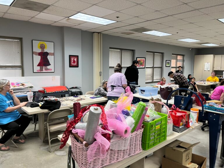 A wide view of the studio, with bins of ribbon, deco mesh and craft supplies in the foreground and artists working at tables further back.