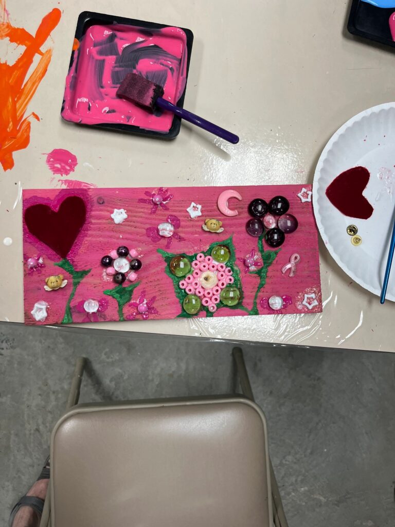 A pink-painted board decorated with a red heart, painted green stems and leaves, and flowers made from glass gems, rhinestone florets and small charms including a crescent moon and a ribbon pin.