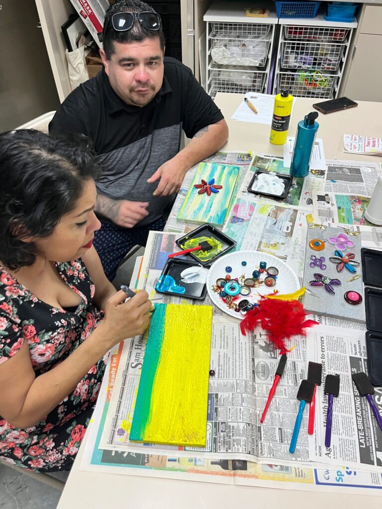 A volunteer helps an artist paint a wooden board in blue and green, with a plate of decorations, feathers and paint trays spread across the newspaper-covered table.