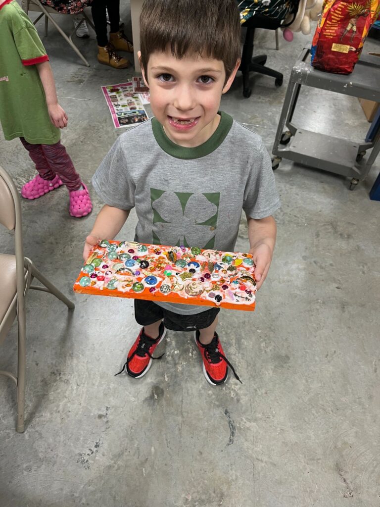 An artist holds up a finished board decorated with a dense cluster of glass gems in blue, green and pink at the centre.