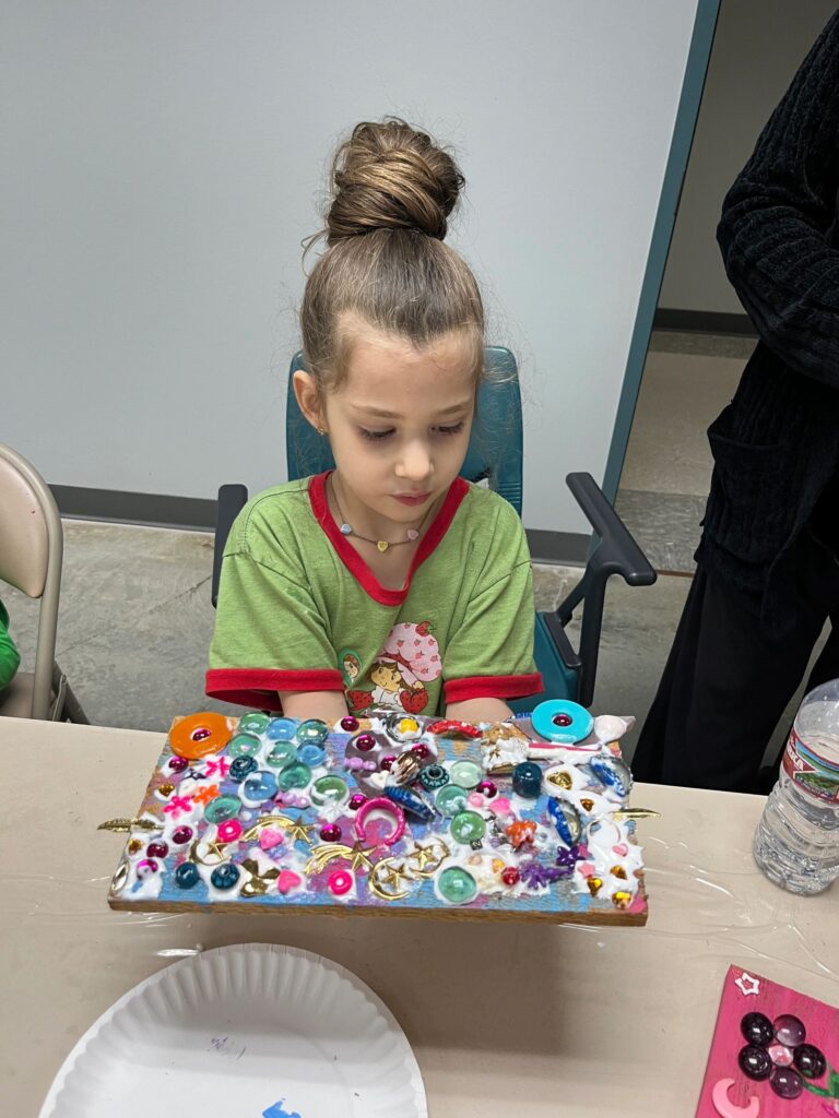 An artist holds a finished board decorated with bottle caps, shells and feather charms in pink, purple and blue, arranged as a flower against a graduated background.