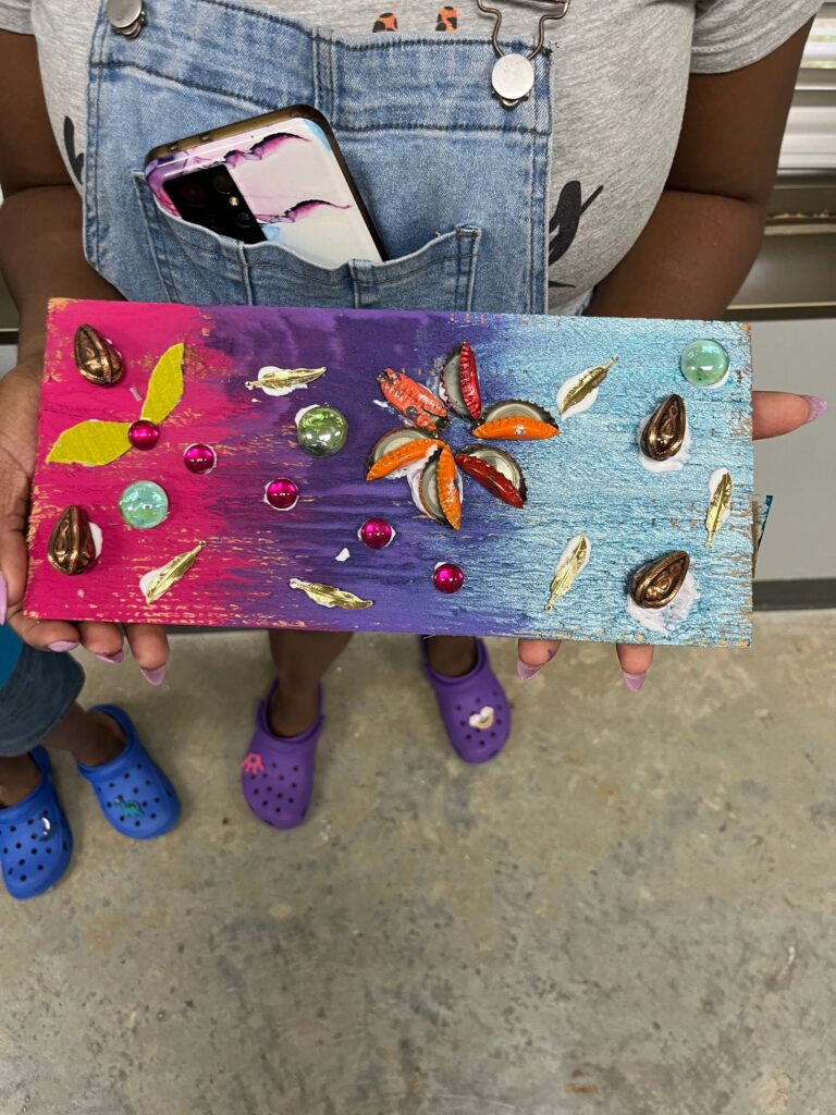 Two artists and a volunteer each hold up their finished boards, decorated with bottle caps, gems and painted flowers in green, purple and orange.