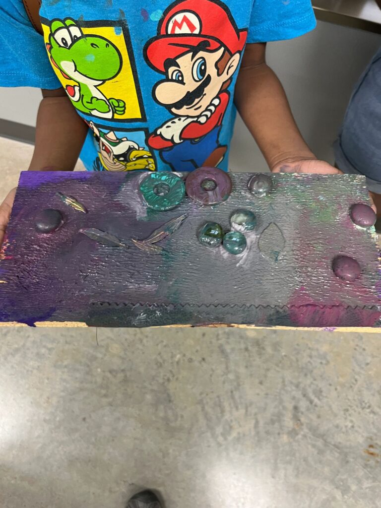 An artist holds a board painted in shades of purple and grey, decorated with glass gems, a metal ring charm and feather charms.