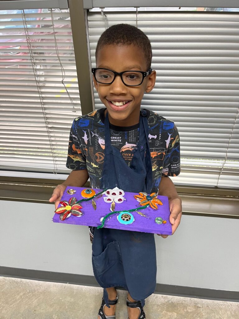 An artist holds a finished purple board decorated with a flower made from painted metal charms, gems and a beaded vine connecting two flower shapes.