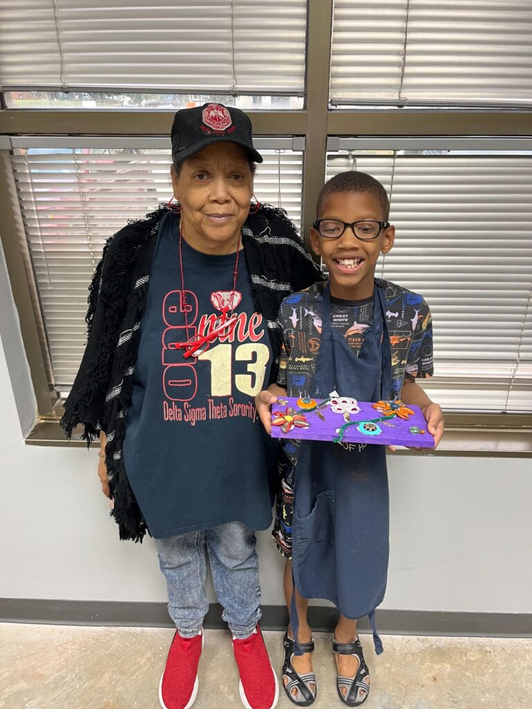 An artist and a volunteer stand together, the artist holding a finished purple board decorated with a flower design.