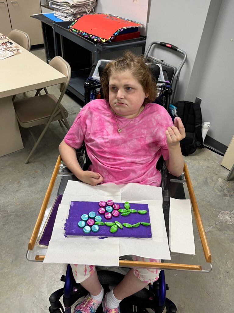 An artist sits with a finished board resting on a wheelchair tray, decorated with bottle caps arranged as a flower in green and pink.