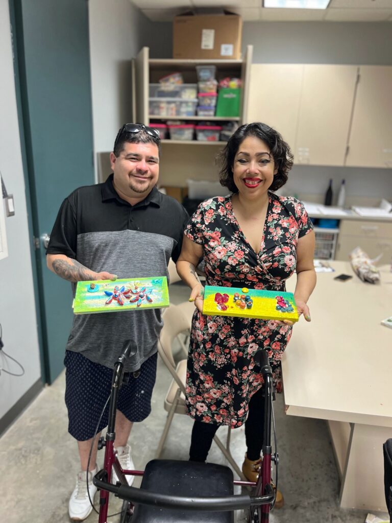 Two artists hold up their finished boards side by side, one decorated with butterfly shapes on a green background, the other with flowers made from bottle caps and gems on a yellow background.
