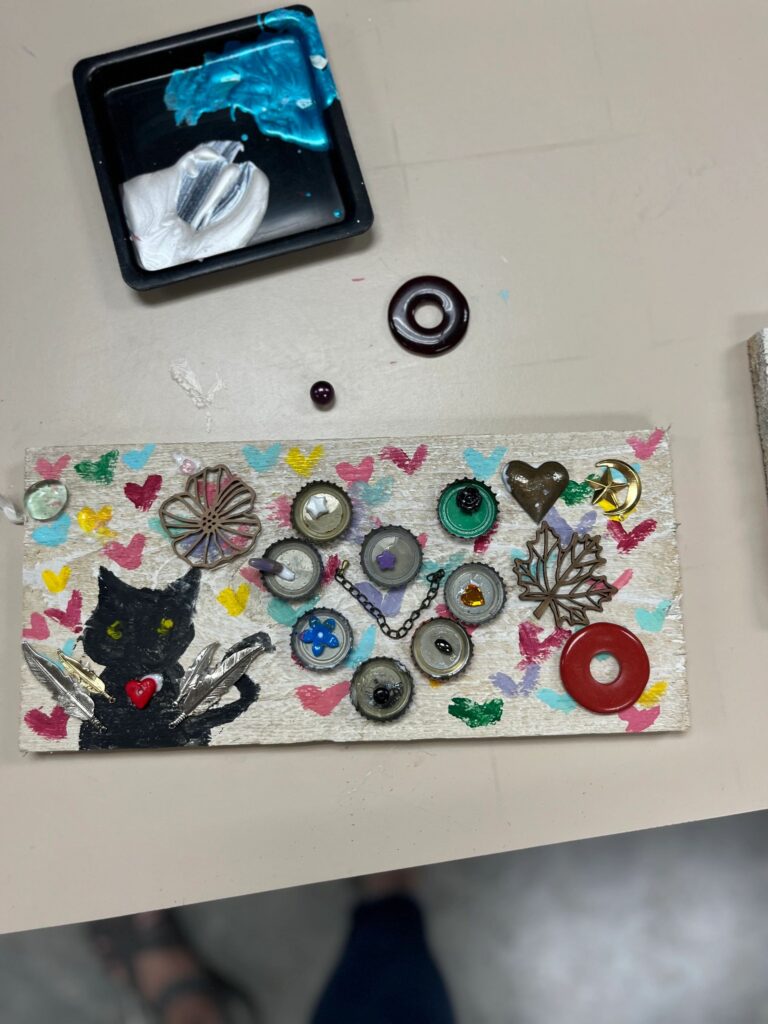 A cream-painted board with a black cat silhouette and hand-painted hearts, decorated with bottle caps, a heart charm, a maple leaf charm and a red donut-shaped bead.