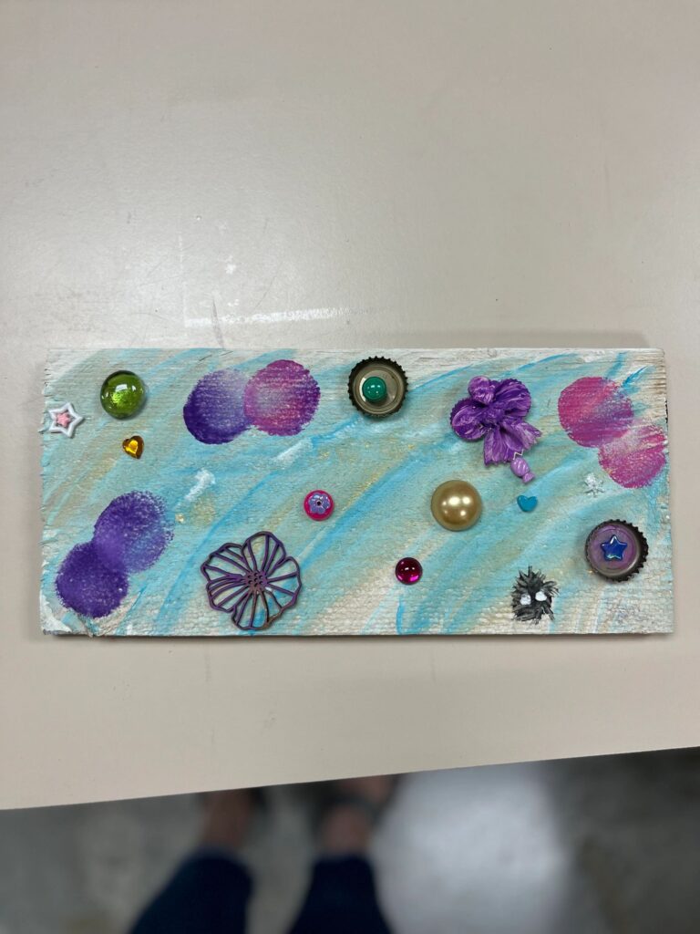 A pale blue and purple board decorated with glass gems, a bottle cap, a fabric flower and a small heart charm, painted with soft cloud-like brushstrokes.