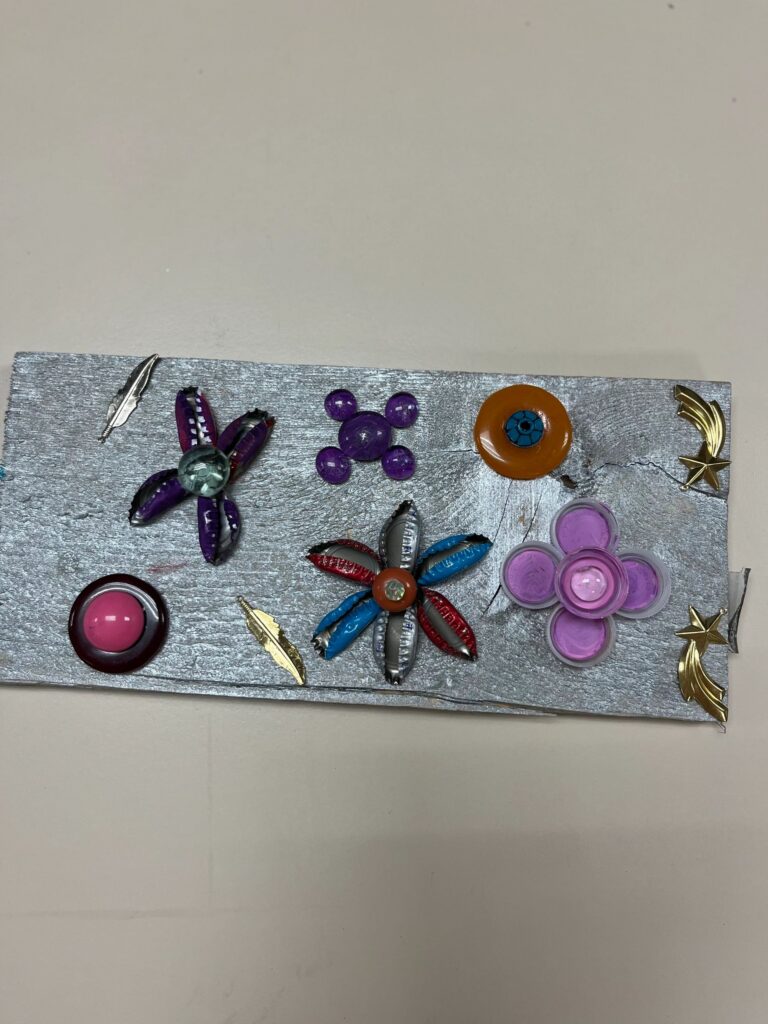 A silver-painted board decorated with flowers made from shell-shaped charms, glass gems and gold star charms arranged in a symmetrical pattern.