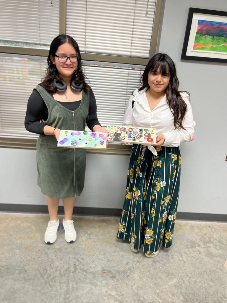 Two artists hold up their finished boards together: one with a pastel cloud design and gem flowers, the other with a black cat silhouette and bottle cap flowers.
