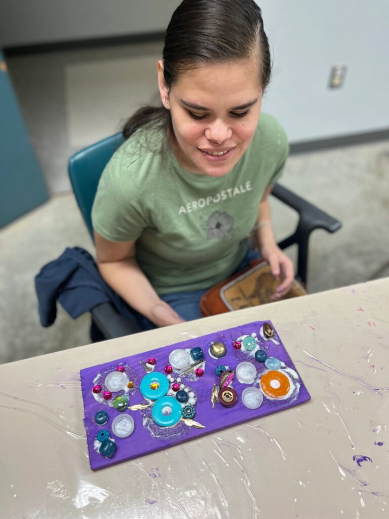 An artist sits with a finished purple board resting on the table, decorated with buttons, glass gems and small charms in teal, orange and pink.