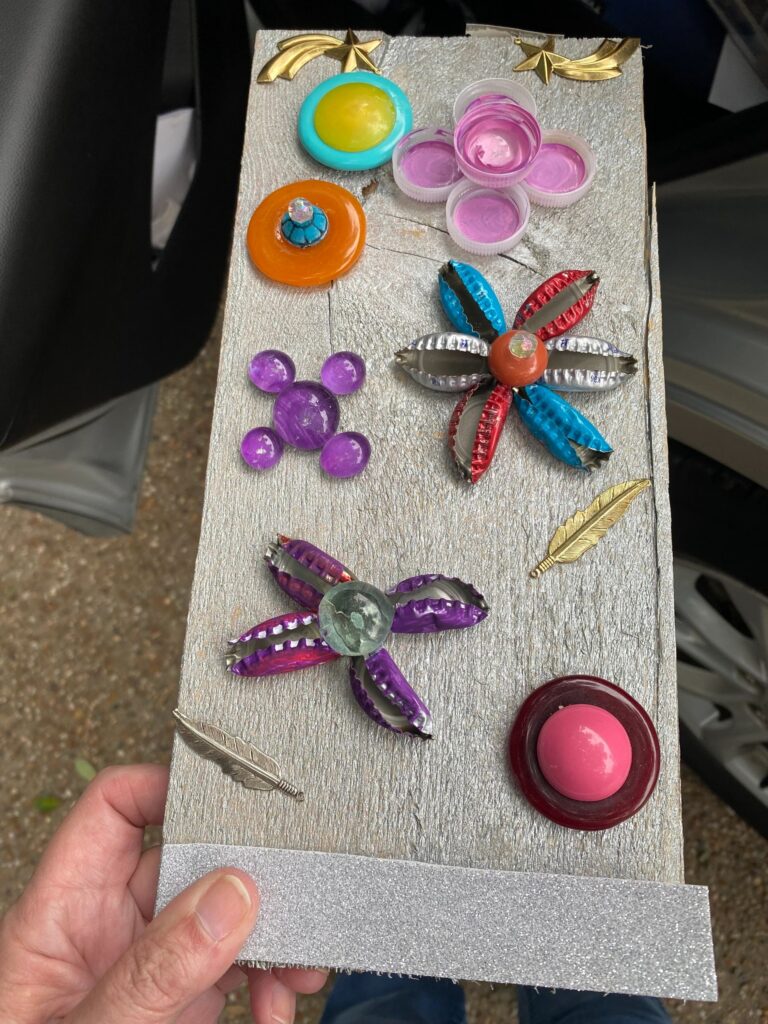 A hand holds a pale wooden board covered in raised, touchable shapes: flowers built from flattened bottle caps in red, blue and purple, clusters of glass marbles, buttons, and small brass feathers and stars.