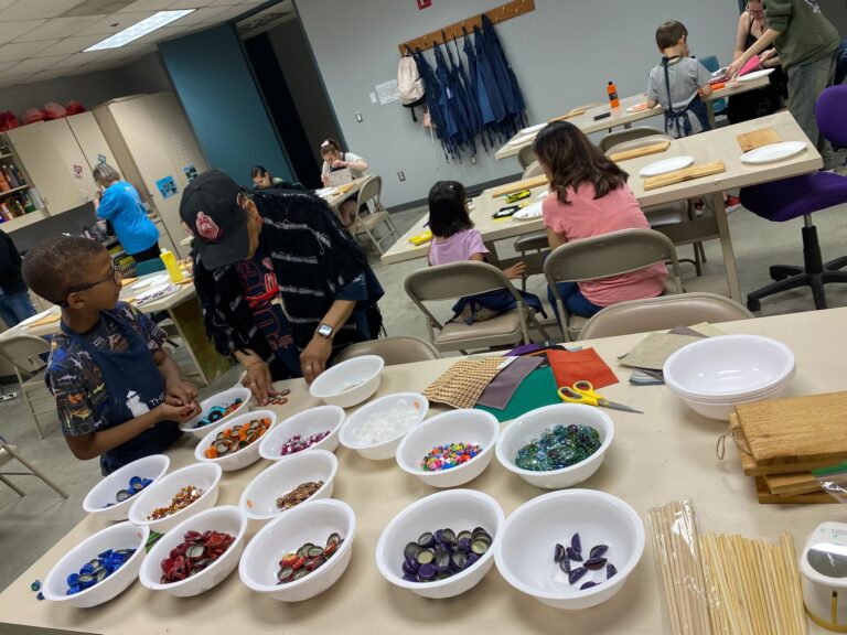 A worktable with bowls of bottle caps, beads and glass gems sorted by colour, plus scissors, ribbon and craft sticks, while artists work on boards in the background.