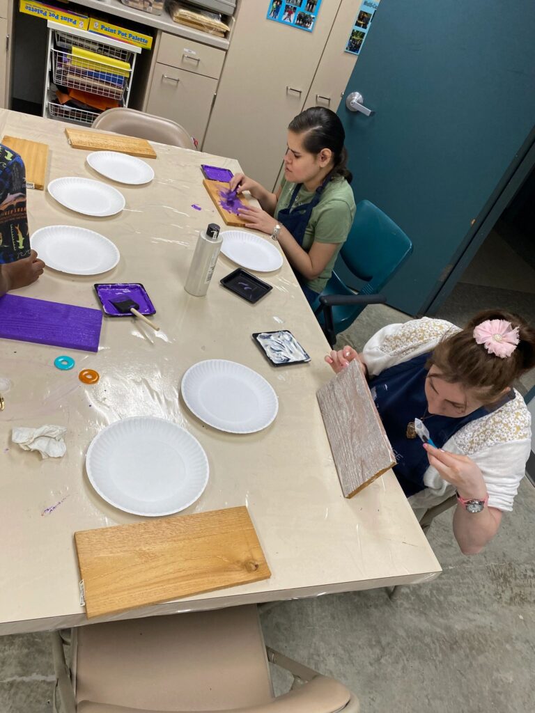 An artist paints a wooden board yellow at a table with paint bottles and paper plates, while others work on boards further down the table.