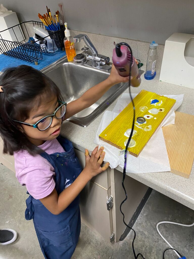 An artist uses a small heat tool to dry a board freshly painted yellow and dotted with bottle caps and glass gems, at a sink counter set up as a drying station.