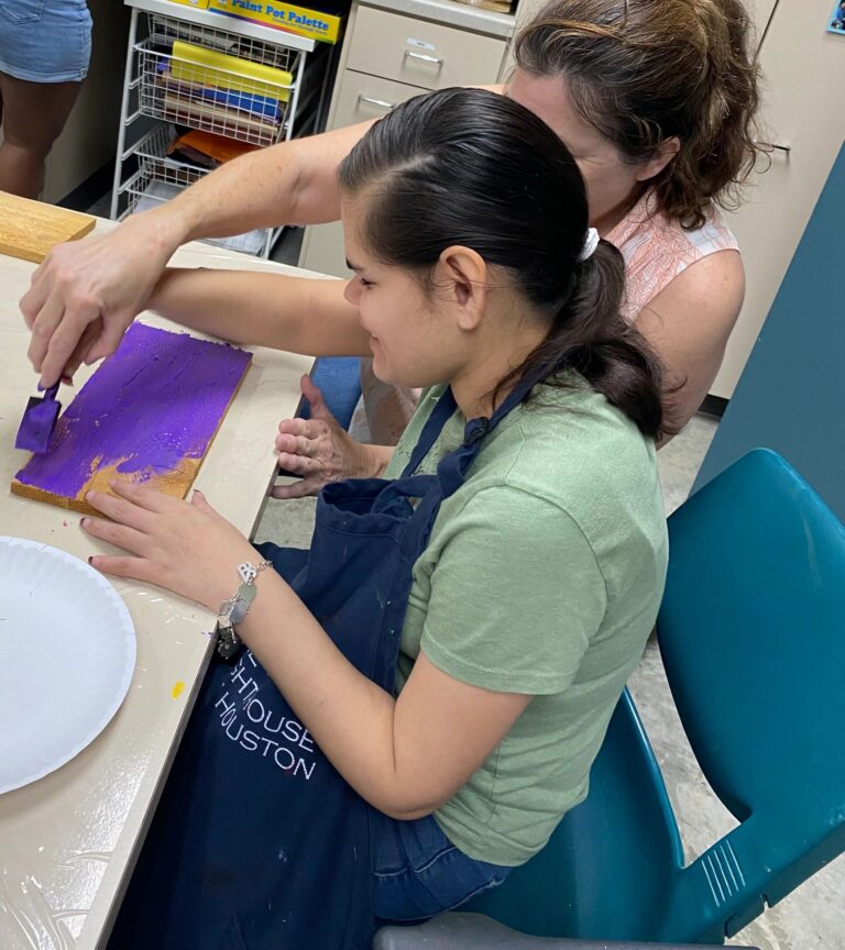 A volunteer helps an artist apply purple paint to a wooden board with a foam brush.