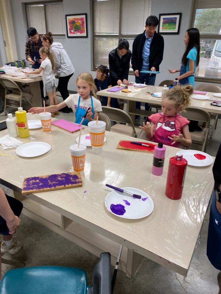 Artists at a table painting and decorating boards with beads and glass gems, with cups, paint bottles and paper plates spread across the tabletop.