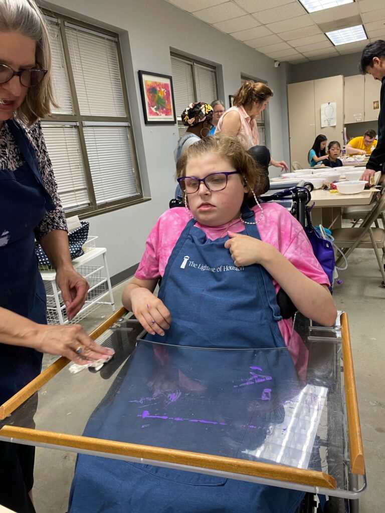 A volunteer helps an artist trace lines with paint on a clear plastic tray fitted to their wheelchair, working alongside other volunteers at nearby tables.