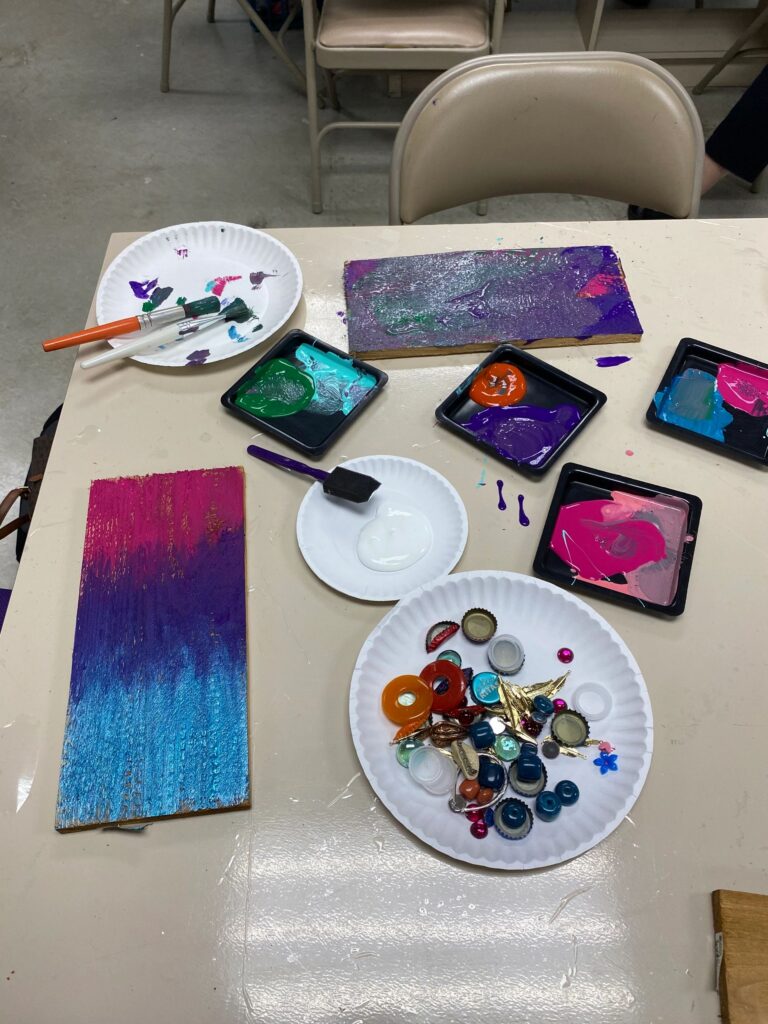 A close-up of art supplies on a table: two painted boards, palettes of blue, purple, pink and orange paint, and a plate of bottle caps and glass gems ready to be added.