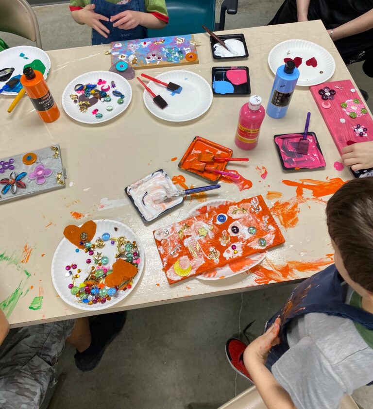 A table covered in painted boards and paint palettes in orange, pink and blue, with beads, gems and bottle caps set out for decorating.