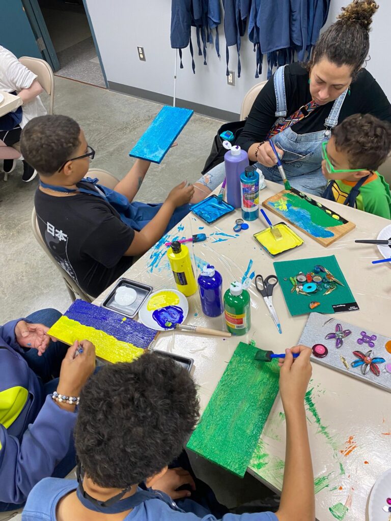 A group of artists at a table painting boards in blue, green, purple and yellow, with paint bottles, trays and a decorated board with pressed flowers in the mix.