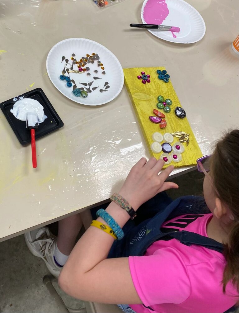 An artist presses beads, gems and small metal shapes into wet paint on a yellow-painted board, with a plate of extra materials nearby.