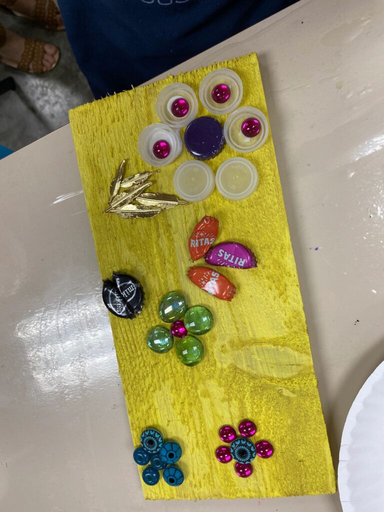 A finished yellow board decorated with bottle caps, glass gems and a gold feather charm, arranged in a loose cluster near the top.