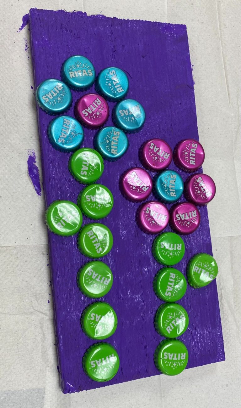 A finished purple board covered in bottle caps arranged as two flower shapes, one in teal, magenta and green, the other in green alone.