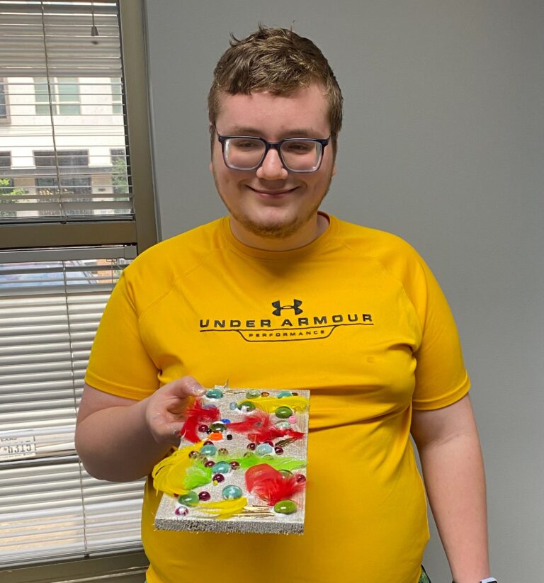 An artist holds up a finished board covered in a dense cluster of glass gems and feathers in red, green and yellow.
